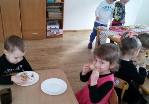 Maluszki siedzą przy stolikach i jedzą śniadanie. W tle widać dwóch chłopców, którzy stoją przy stoliku i przygotowują sobie kanapki.
