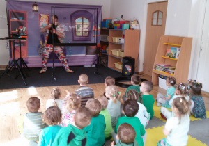 Widok na dzieci siedzące na dywanie oraz na Pana Bańkę prezentującego pokaz baniek mydlanych.