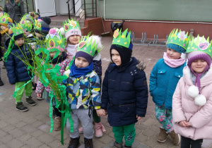 Widok na grupę dzieci, które mają na głowach papierowe zielone opaski z motylkami, biedronkami, pszczółkami i kwiatkami. Fabian trzyma w ręku gałązki przystrojone zieloną bibułą.