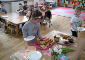 Widok na grupę siedzących przy stolikach dzieci. Laura i Oluś przygotowują sobie kanapki korzystając ze "szwedzkiego stołu".