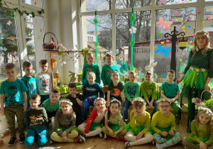 Dzieci w strojach zielonych pozują do zdjęcia grupowego z Panią wiosna
