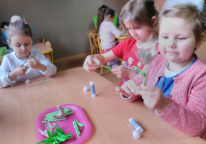 Dziewczynki siedzące przy stoliku przymocowują rurki z papieru do kielicha tulipana, przyklejają listki.