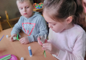 Widok na dwoje dzieci siedzących przy stoliku, dziewczynka przymocowuje rurkę z papieru do kielicha tulipana, przykleja listki.