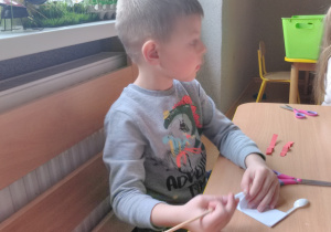 Chłopiec siedząc przy stoliku skleja elementy bociana. Na stole miseczka z klejem, pędzel i nożyczki.