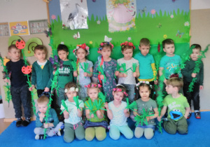 Grupa dzieci pozuje do zdjęcia, stojąc na tle tablicy udekorowanej w wiosenną łąkę. Dziewczynki mają na głowie wianki, chłopcy trzymają w rękach gałązki zielone z symbolami wiosennymi.
