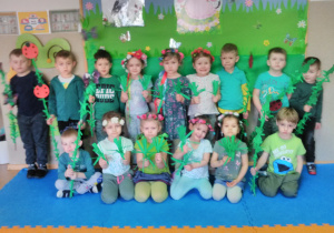 Grupa dzieci pozuje do zdjęcia, stojąc na tle tablicy udekorowanej w wiosenną łąkę. Dziewczynki mają na głowie wianki, chłopcy trzymają w rękach gałązki zielone z symbolami wiosennymi.