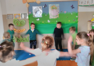 Dzieci stojąc w kole ilustrują ruchem piosenkę „Wiosna wita nas”. W tle tablica z dekoracją wiosenną.