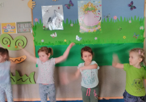 Dzieci stojąc w kole ilustrują ruchem piosenkę „Wiosna wita nas”. W tle tablica z dekoracją wiosenną.