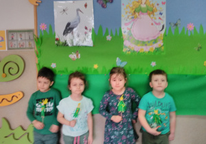 Dzieci na tle tablicy udekorowanej w łąkę wiosenną prezentują swoje wytwory plastyczne – kukiełkę Pani Wiosny.