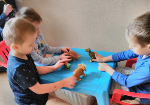 Chłopcy siedząc przy małym, niebieskim stoliku bawią się figurkami dinozaurów.
