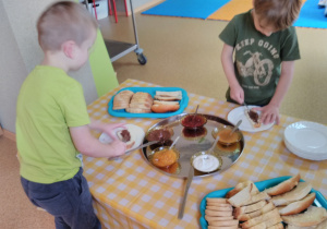 Dzieci stoją przy stole nakrytym obrusem w żółtą kratę. Na stole taca srebrna z miseczkami, z dżemem truskawkowym i brzoskwiniowym, masłem orzechowym, miodem i serkiem waniliowym. Na drugiej tacy srebrnej pieczywo: rogaliki, angielka, chałka. Dzieci smarują pieczywo masłem orzechowym.