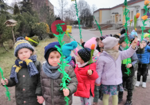Dzieci stoją parami na placu przedszkolnym z rekwizytami w dłoni (gałązki zielone z biedronkami, żabkami, motylami, kwiatkami).