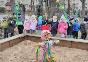 Dzieci stoją wokół piaskownicy. Żegnają kukłę „Pani Zimy” umieszczoną w środku piaskownicy. W rękach trzymają zielone gałązki.