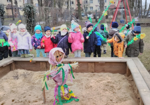 Dzieci stoją wokół piaskownicy. Żegnają kukłę „Pani Zimy” umieszczoną w środku piaskownicy. W rękach trzymają zielone gałązki.