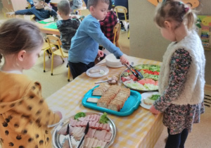 Dzieci stoją przy stole nakrytym obrusem w kratę: biało-żółtą. Na stole tace z nowalijkami, pieczywem oraz wędliną. Chłopiec chwyta szczypcami plastry zielonego ogórka.