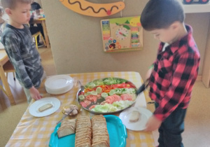 Dwóch chłopców stoi przy stole nakrytym obrusem w kratę: biało-żółtą. Na stole tace z nowalijkami, pieczywem oraz wędliną. Chłopiec w prawej ręce trzyma szczypce i chwyta plastry pomidora. W drugiej ręce trzyma talerz z kromką chleba.