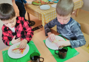 Dwóch chłopców siedzi przy stole i dekoruje kanapki nowalijkami.