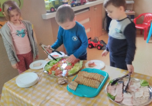 Troje dzieci stoi przy stole nakrytym obrusem w kratę: biało-żółtą. Na stole tace z nowalijkami, pieczywem oraz wędliną. Chłopiec szczypcami chwyta nowalijki: ogórki, sałatę.