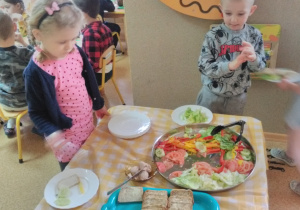 Dwoje dzieci stoi przy stole nakrytym obrusem w kratę: biało-żółtą. Na stole tace z nowalijkami, pieczywem oraz wędliną. Dzieci patrzą na tace z nowalijkami i dokonują wyboru warzywa na kanapkę.