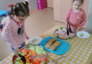 Emilka i Maja stoją przy stole nakrytym obrusem w kratę: biało-żółtą. Na stole tace z nowalijkami, pieczywem oraz wędliną. Dziewczynki szczypcami chwytają paprykę i wędlinę.
