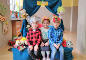 Troje dzieci pozuje do zdjęcia w czapkach bajkowych: myszki, koguta, kaczki. W tle teatrzyk z zieloną kurtyną i napisem „TEATR”.