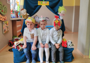 Troje dzieci pozuje do zdjęcia w czapkach bajkowych: myszki, koguta, kaczki. W tle teatrzyk z zieloną kurtyną i napisem „TEATR”.