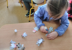 Dziewczynka siedzi przy stoliku, składa postacie bajkowe zrobione z papieru i skleja ich boki