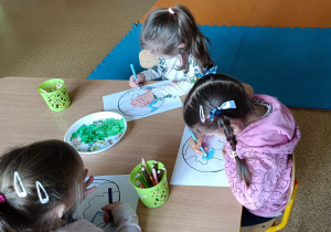 Troje dzieci siedzi przy stole i malują kontury Ziemi zieloną kredką.