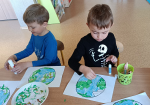 Michał i Kacper siedzą przy stoliku i wyklejają kontury Ziemi zielona bibułą.