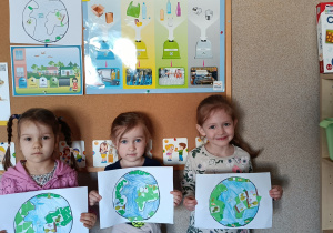 Prezentacja prac plastycznych „Planeta Ziemia” przez 3 dziewczynki stojące na tle tablicy z obrazkami recyklingu.