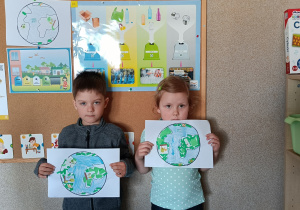 Prezentacja prac plastycznych „Planeta Ziemia” przez 2 dzieci stojących na tle tablicy z obrazkami recyklingu.