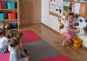Widok na Laurę, która siedzi na krzesełku przed tablicą z papierową krową i demonstruje dzieciom jej dojenie.