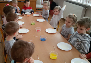 Widok na dzieci siedzące przy zestawionych razem stolikach. Przed nimi stoją talerzyki z mlekiem i pojemniki z farbami.