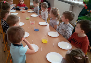 Widok na dzieci siedzące przy stoliku. Przed nimi stoją talerzyki z mlekiem i pojemniki z farbami.