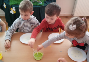 Widok na troje dzieci, które moczą patyczki higieniczne w zielonej farbie.