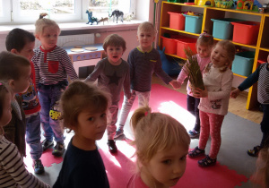 Przedszkolaki tańczą w kole przy piosence. W środku koła stoi Zuzia, która trzyma w rękach roślinkę.