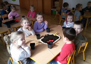 Widok na dzieci siedzące przy stolikach, na których znajdują się tace z ziemią łopatki, małe plastikowe doniczki.