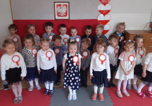 Dzieci ubrane w stroje galowe, z przypiętymi kokardami narodowymi pozują do zdjęcia grupowego na tle biało- czerwonej tablicy.