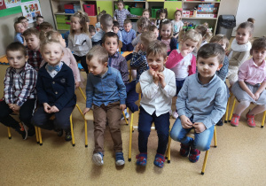 Widok na grupę siedzących na krzesłach przedszkolaków.