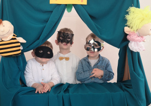 Widok na trzech chłopców w ozdobnych maskach, którzy stoją w okienku teatrzyku przedszkolnego.