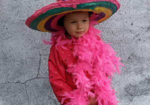 Widok na dziewczynkę, która pozuje do zdjęcia w szalu z różowych piór i dużym kolorowym kapeluszu.