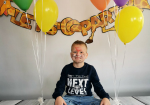Chłopiec siedzi na tle fotograficznym i pozuje do zdjęcia wśród balonów.