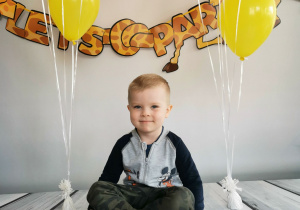 Chłopiec siedzi na tle fotograficznym i pozuje do zdjęcia wśród balonów.
