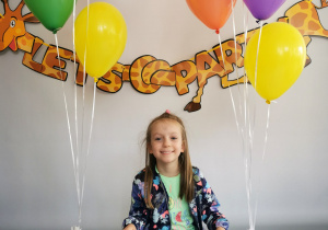 Dziewczynka siedzi na tle fotograficznym i pozuje do zdjęcia wśród balonów