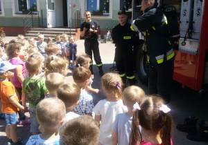 Widok na grupę dzieci i trzech Strażaków prezentujących dzieciom sprzęt strażacki.