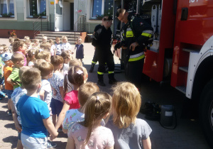 Widok na grupę dzieci i trzech Strażaków prezentujących dzieciom sprzęt strażacki.