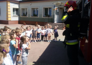 Widok na grupę dzieci i strażaka zakładającego hełm strażacki.