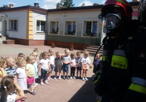 Widok na grupę dzieci i strażaka w specjalnym stroju do gaszenia pożaru.