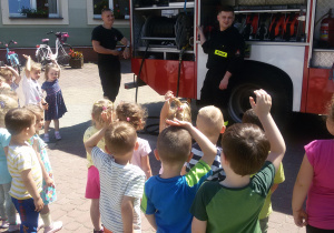 Widok na grupę dzieci, dwóch strażaków i samochód strażacki.