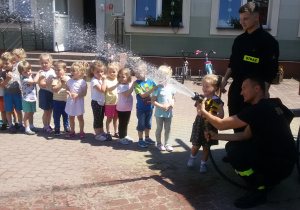 Widok na grupę dzieci i dziewczynkę, która leje wodą z węża strażackiego.